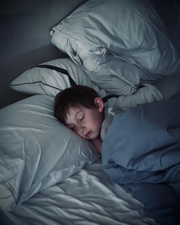 Child sleeps peacefully in bed with white pillows and blue blanket in a dimly lit room at nighttime. Sleeping child in bed.