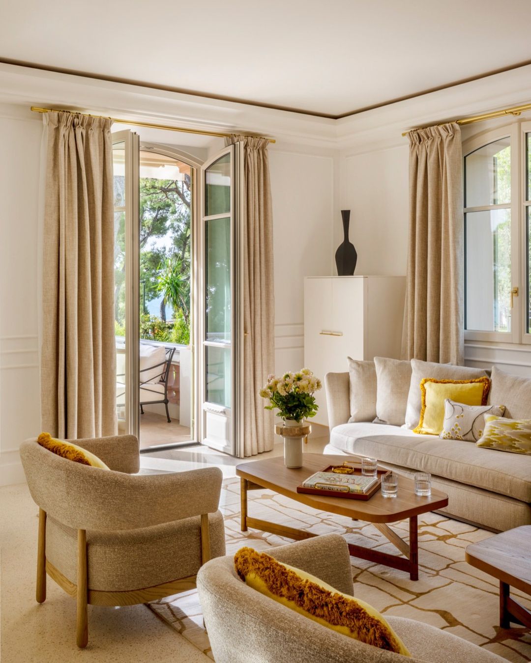 Bright living room with beige armchairs, white sofa, wooden coffee table, and open French doors leading to a balcony.