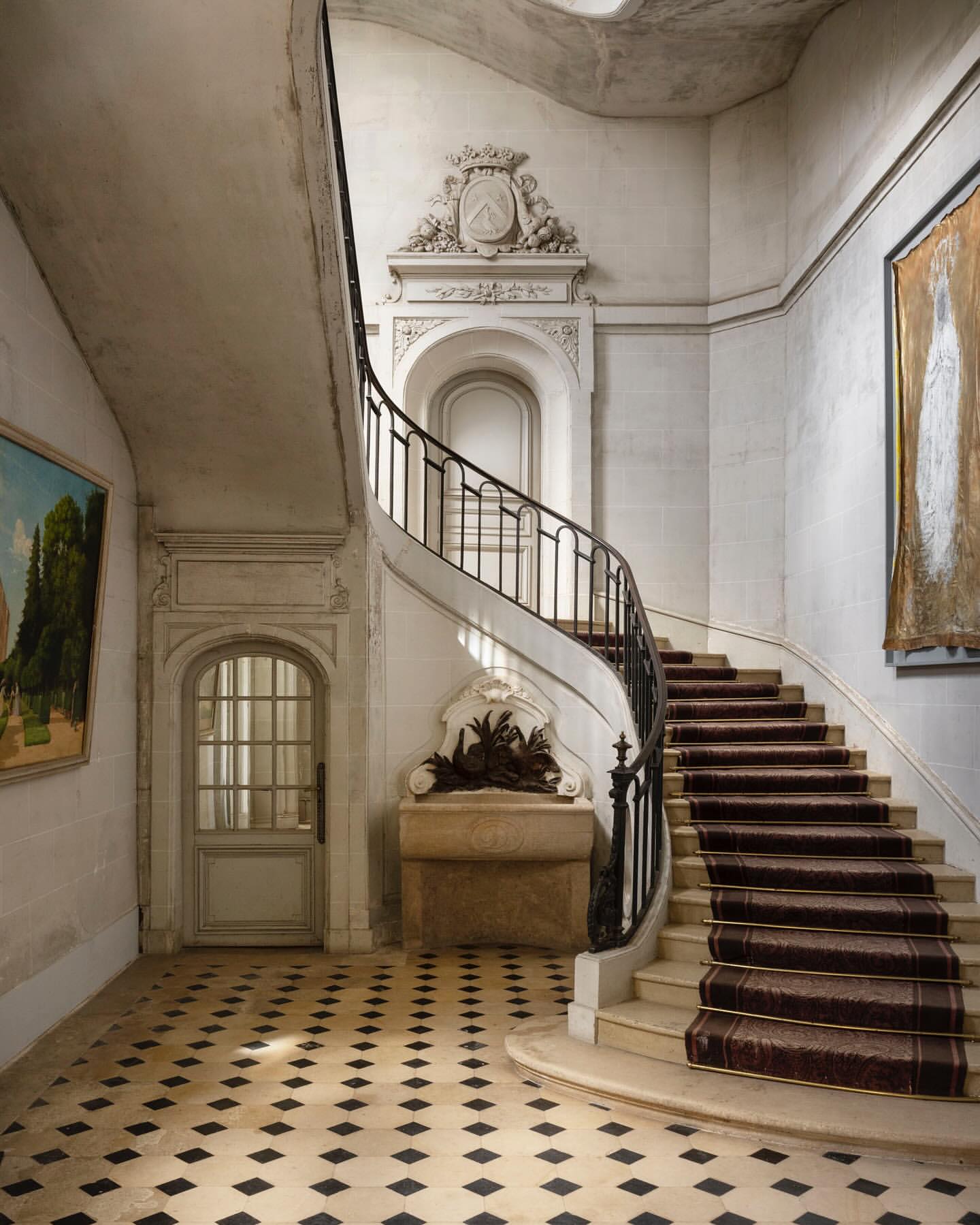 Ornate stairway with brown carpet runner, white marble stairs, stone wall, and artwork inside an elegant building's interior.