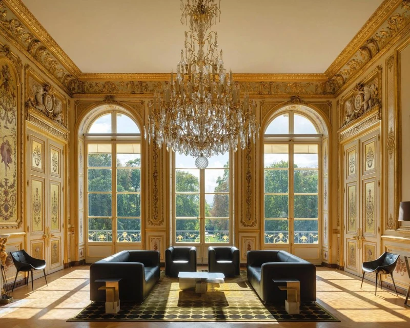 Ornate room with black sofas, chairs, and a chandelier. Natural light streams through the large windows, illuminating the classic interior.