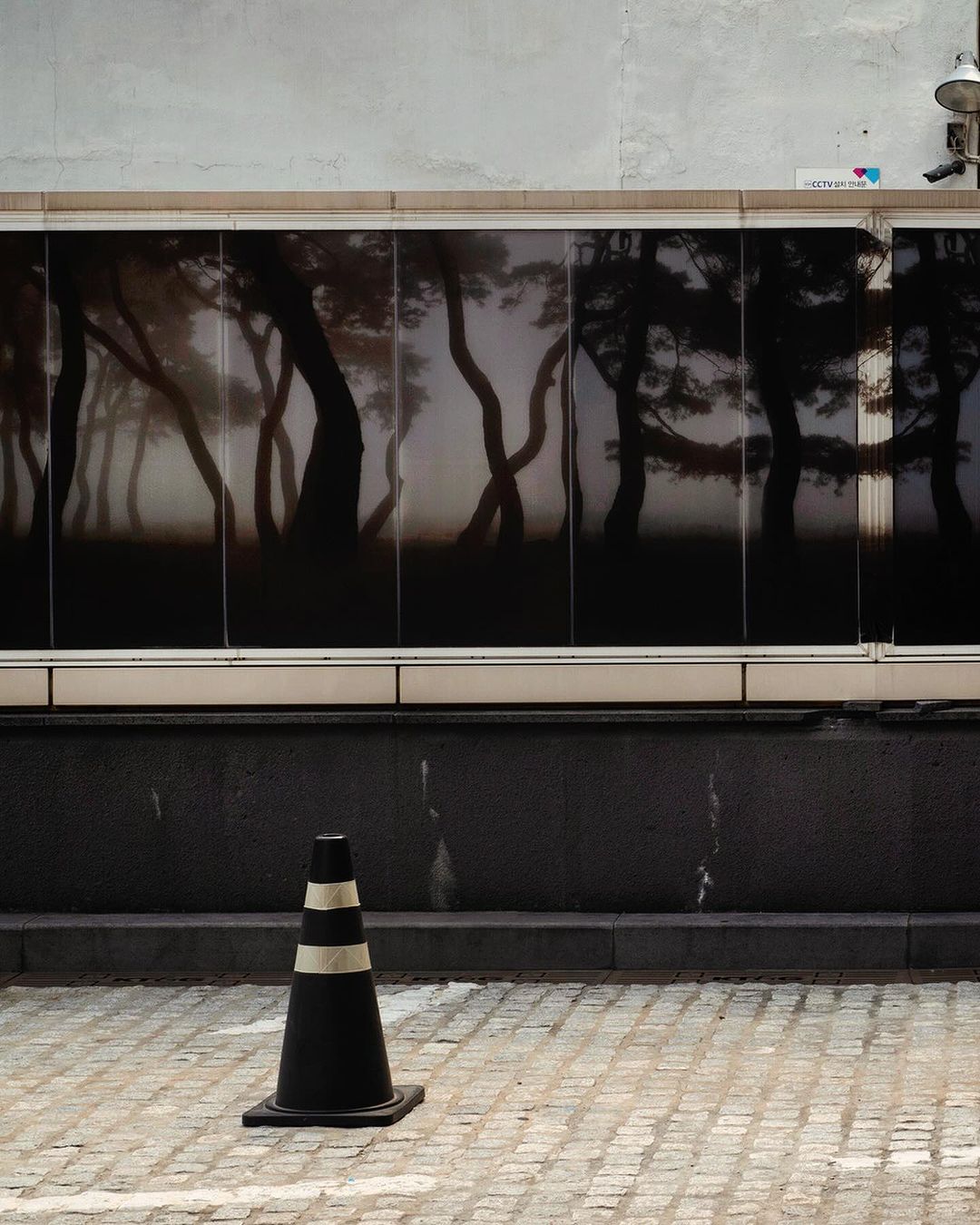 A black and white striped traffic cone sits on a gray brick surface, in front of a large window displaying an abstracted forest image.