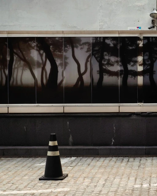 A black and white striped traffic cone sits on a gray brick surface, in front of a large window displaying an abstracted forest image.