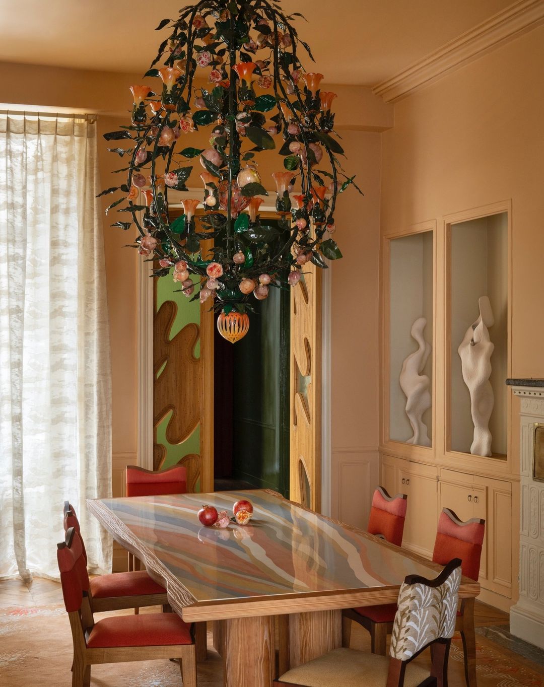 Eclectic dining room interior with a wooden table, red chairs, and a decorative floral chandelier hanging overhead, next to a doorway.