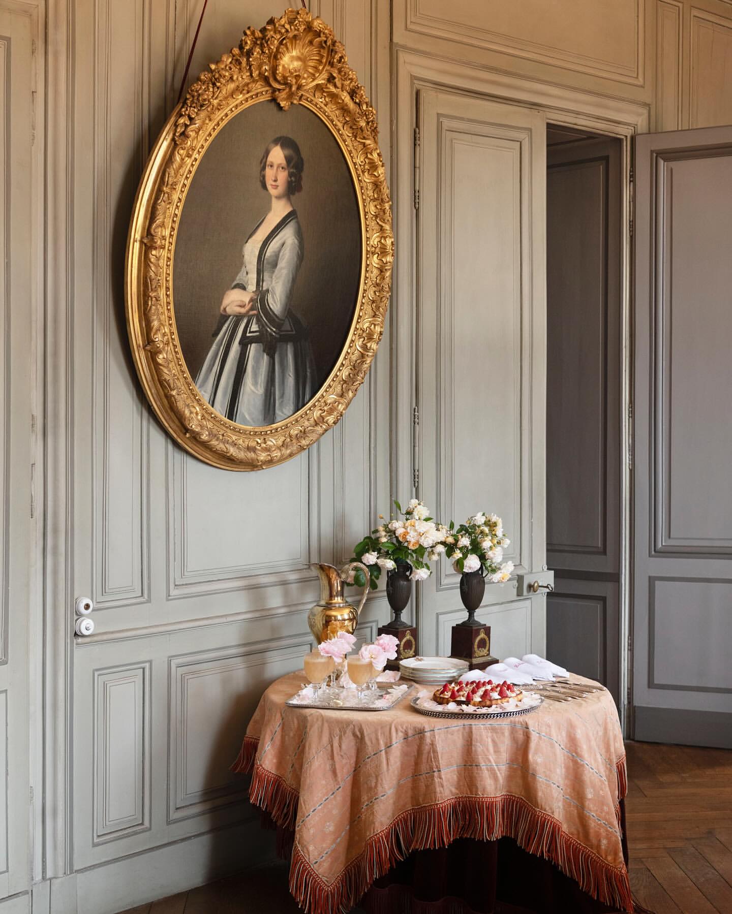 Interior view with a golden-framed portrait hanging on a wall above a table set with drinks, a dessert, flowers and decorative objects.