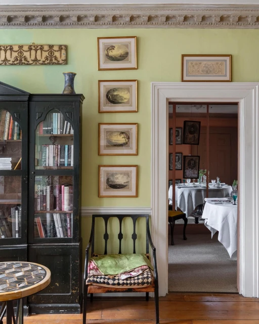Interior of a room featuring a black cabinet filled with books, framed artwork on a light green wall, a chair with cushions, and an open doorway.