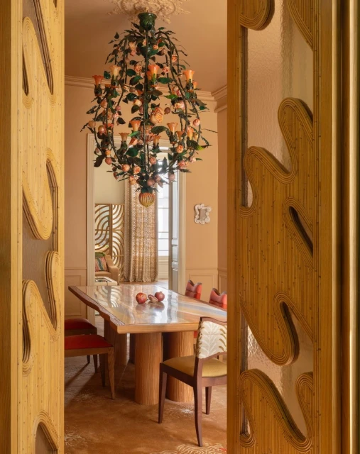 Elegant dining room with an elaborate floral chandelier, dining table with fruit, and carved wood doorway viewed from a doorway.