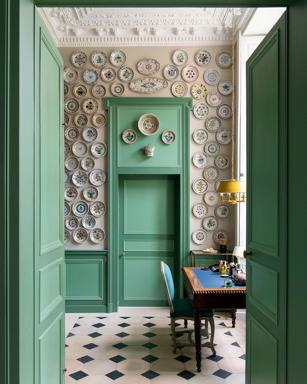 Interior view of a room featuring a wall covered with decorative plates, green-painted doorframes, and a desk visible through doorway.