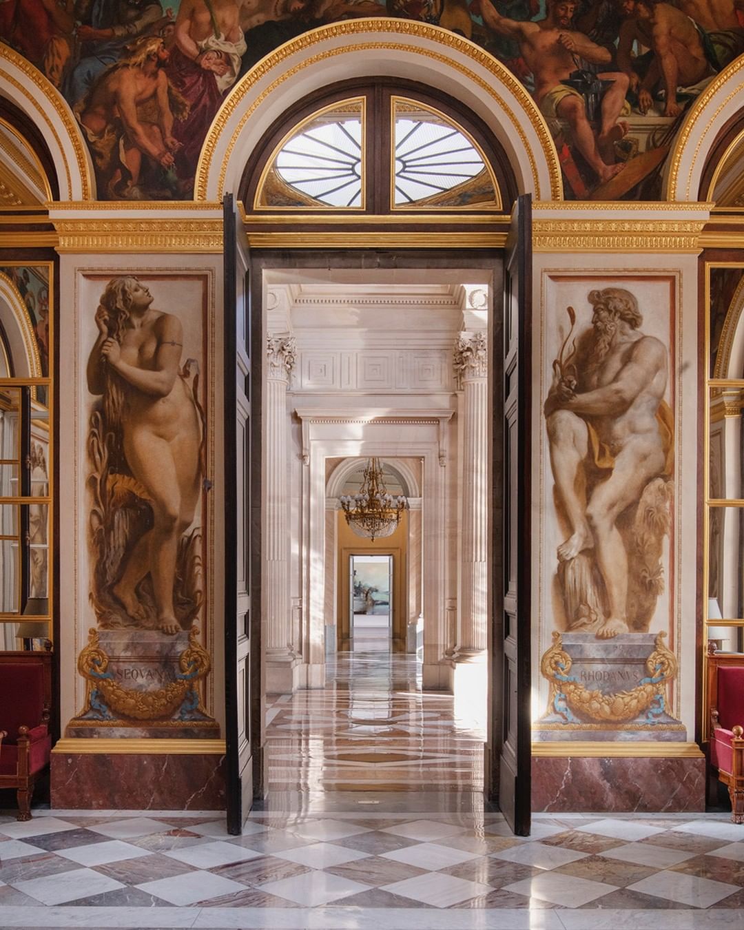 Architectural shot inside a long hallway, adorned with classical statues, marble floors, and mural paintings, evoking a sense of grand elegance.