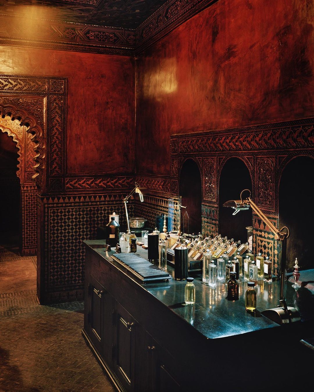 Interior view of a dimly lit room featuring a table laden with perfume bottles, illuminated by desk lamps, against ornate wall decor.