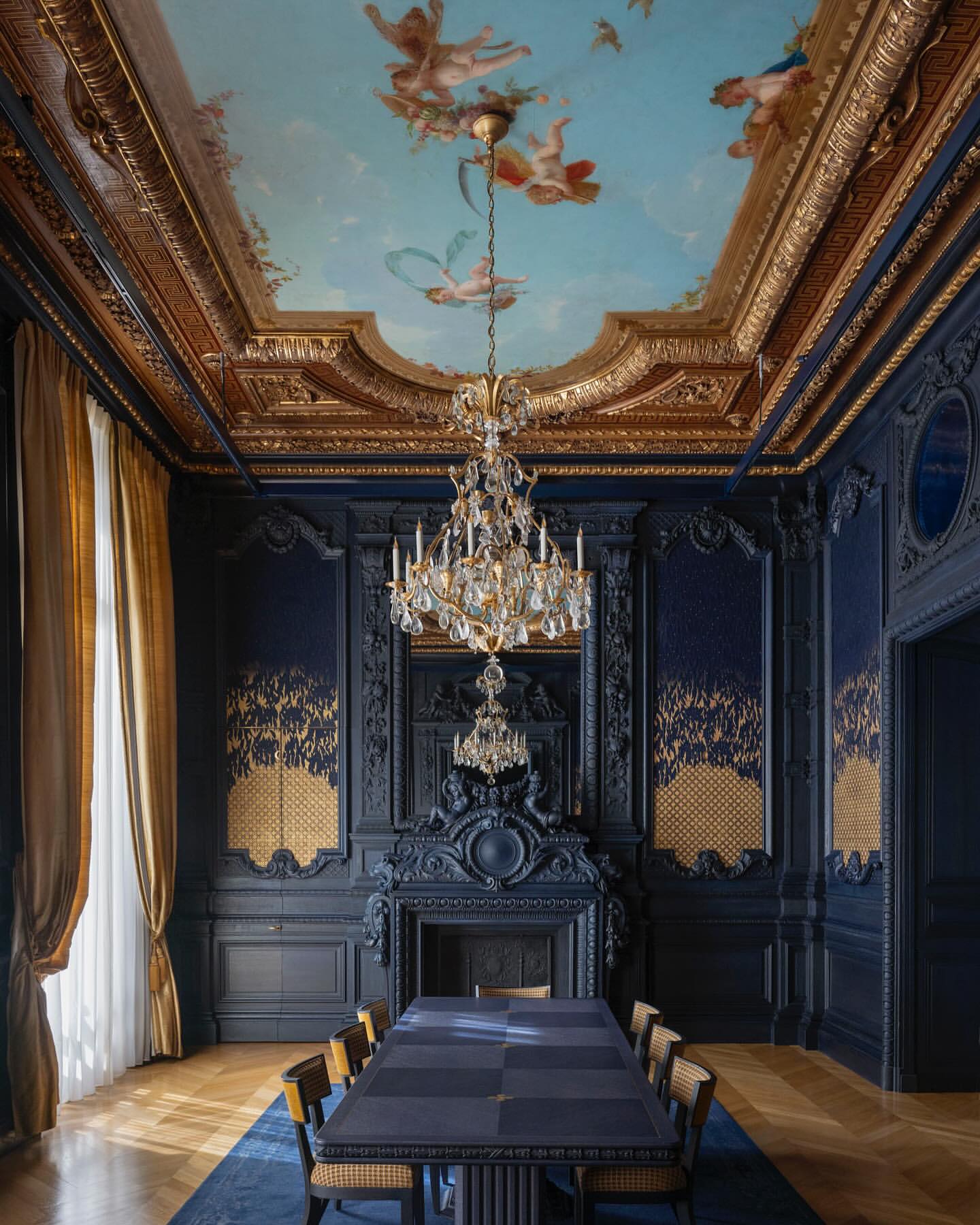 Ornate dining room interior featuring dark blue walls with gold accents, a crystal chandelier, and a painted ceiling with cherubs.