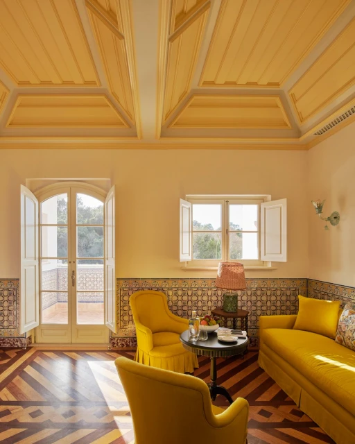 Ornate room featuring a yellow sofa and chairs, a round table with fruit, and open French doors leading to a sunlit balcony.