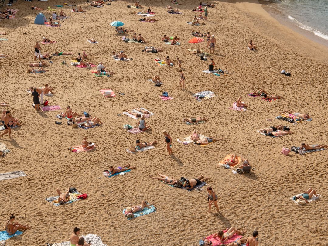 Aerial view of a crowded sandy beach with many people sunbathing, swimming, and relaxing in swimwear during a sunny summer day.