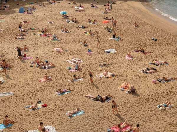 Aerial view of a crowded sandy beach with many people sunbathing, swimming, and relaxing in swimwear during a sunny summer day.