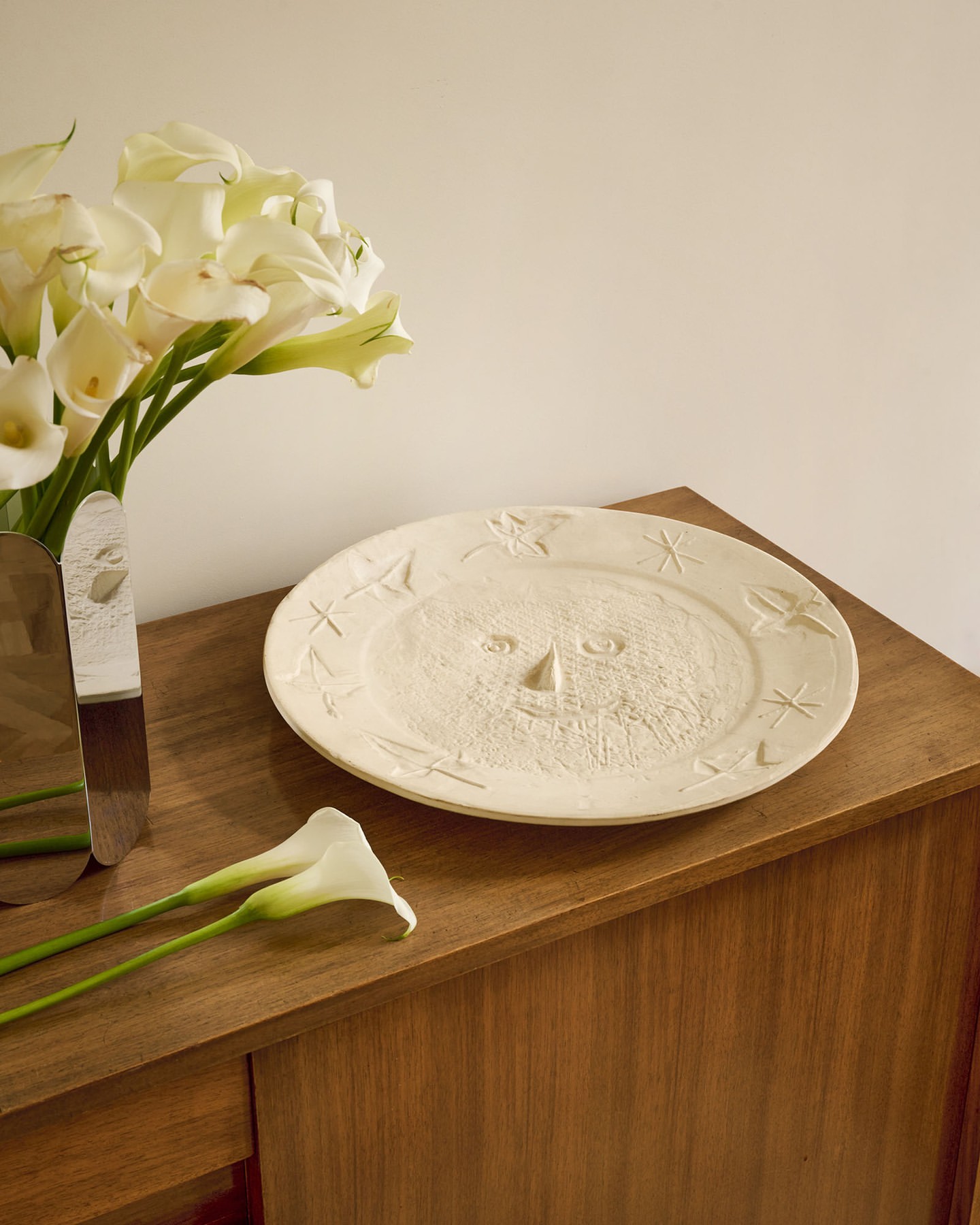 Close-up of a decorative plate with a sun face design on a wooden table next to a bouquet of white calla lilies.