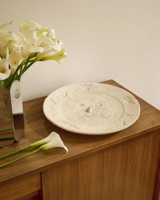 Close-up of a decorative plate with a sun face design on a wooden table next to a bouquet of white calla lilies.