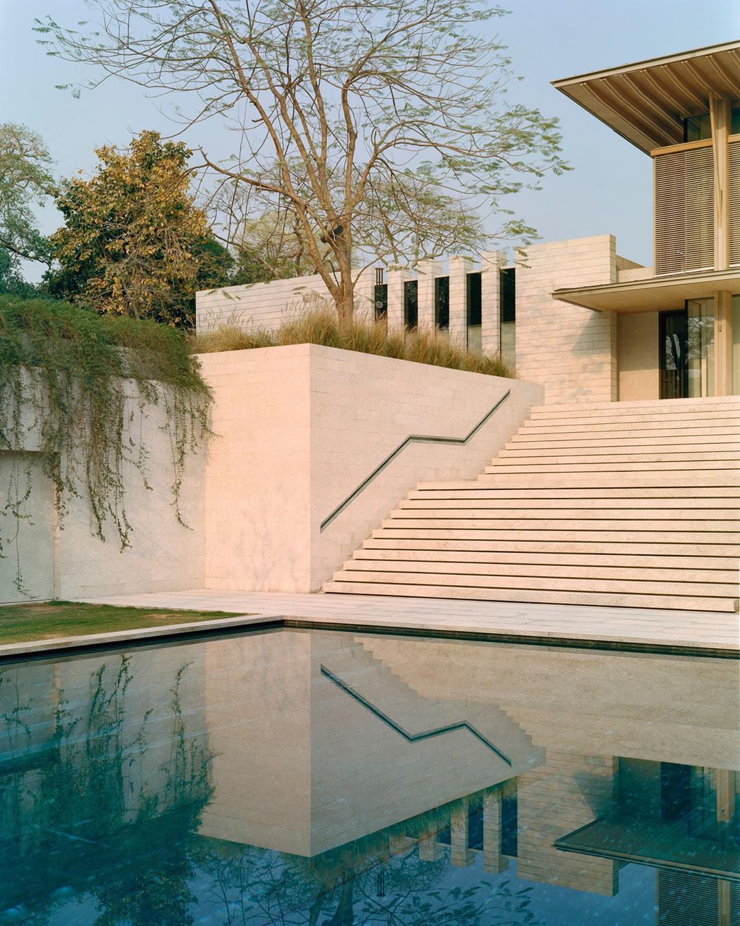 Exterior view of a modern architectural building with travertine walls, a stepped staircase, a reflective pool, and green landscaping.