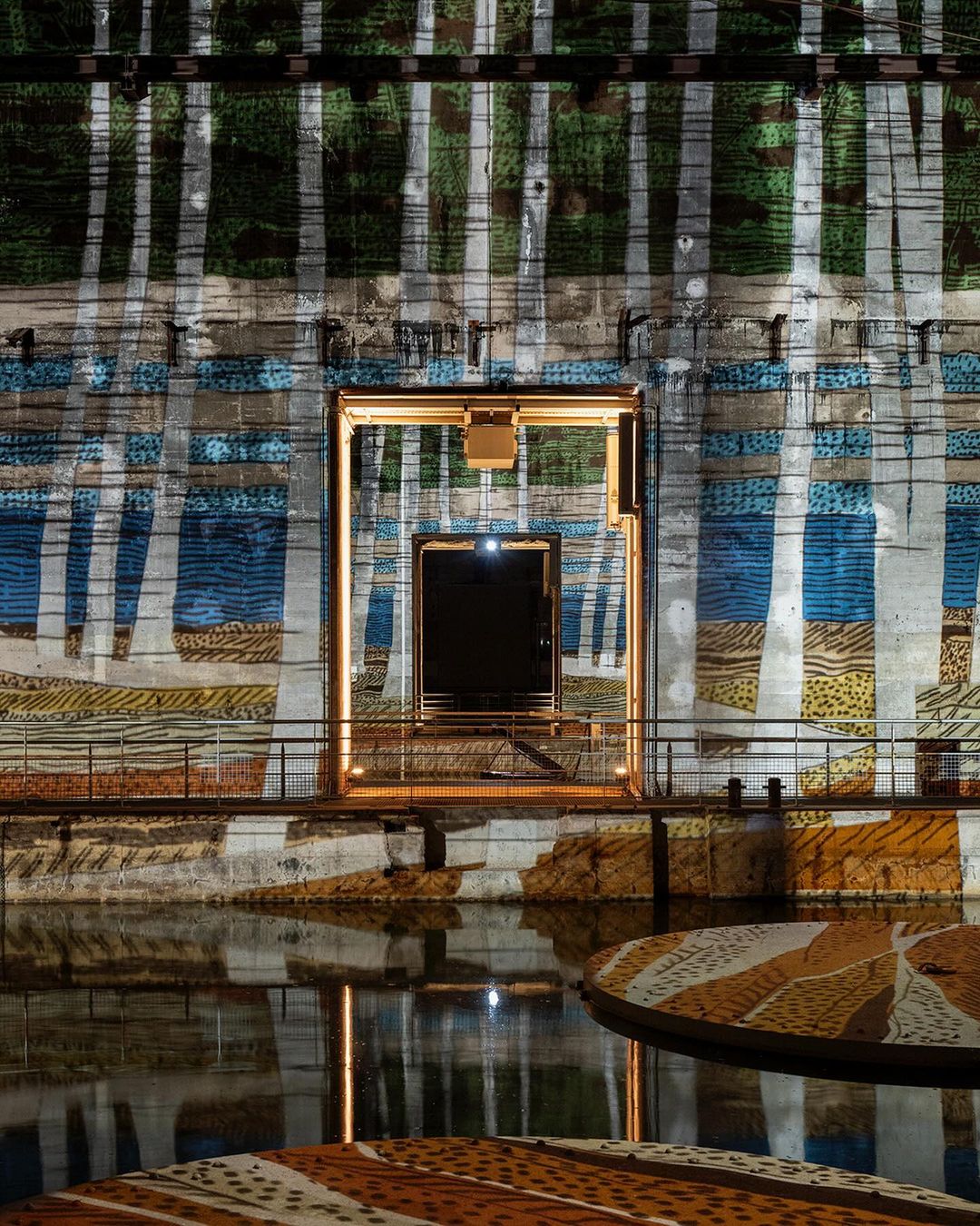 Abstract building facade featuring an open doorway, architectural elements, and lighting reflecting in water, in an urban environment.