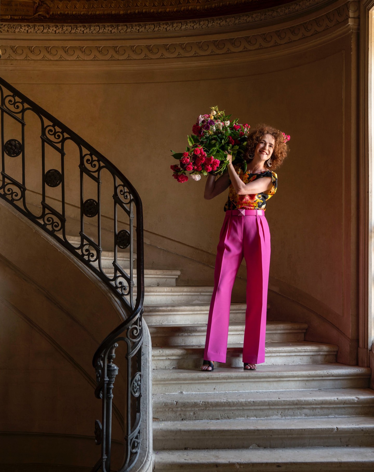 Woman in pink pants holding bouquet on staircase. Floral top, red curly hair, and ornate interior architecture visible.