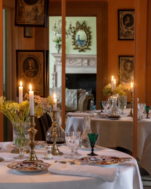 Elegant dining table set with brass candle holders, crystal glassware, floral arrangements, and framed portraits on a warm-toned wall.