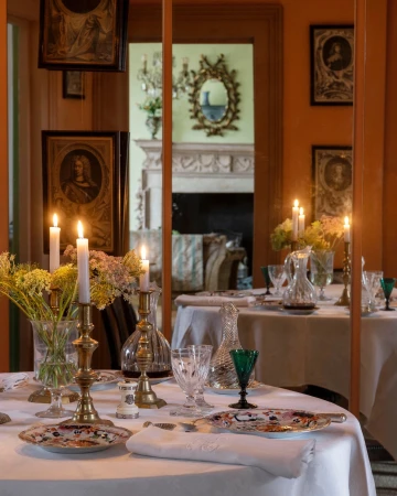 Elegant dining table set with brass candle holders, crystal glassware, floral arrangements, and framed portraits on a warm-toned wall.