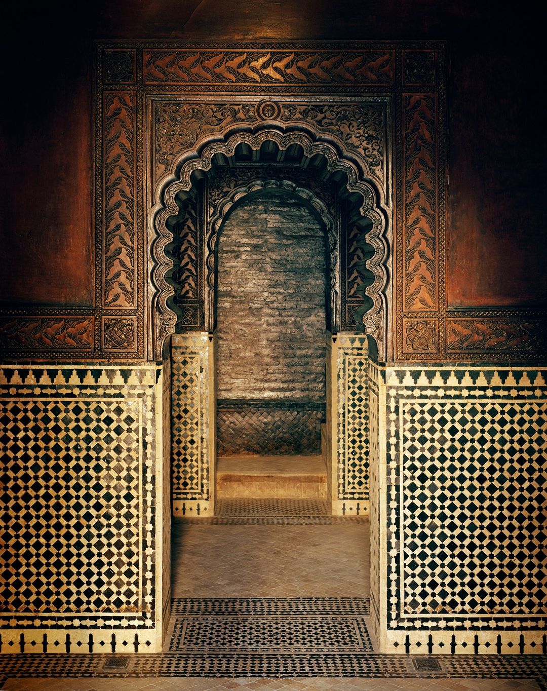 Ornate archway with detailed carvings and tilework in shades of brown, tan, and black, suggesting a Moroccan-style architectural interior.