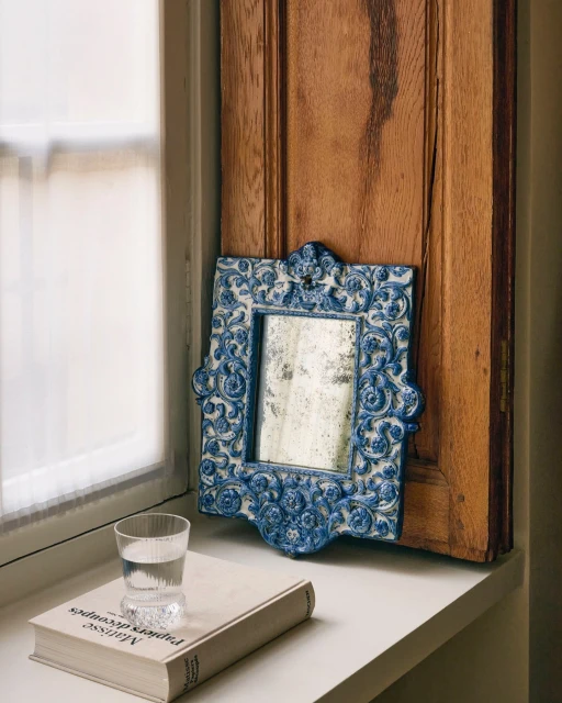 Still life featuring a vintage mirror with a blue floral frame, glass of water, and book on a windowsill with natural light.