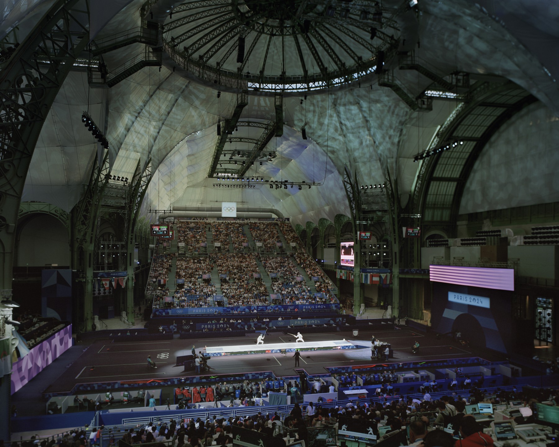 Indoor fencing competition inside an ornate event space filled with spectators, fencing pistes, flags, and large video screens.
