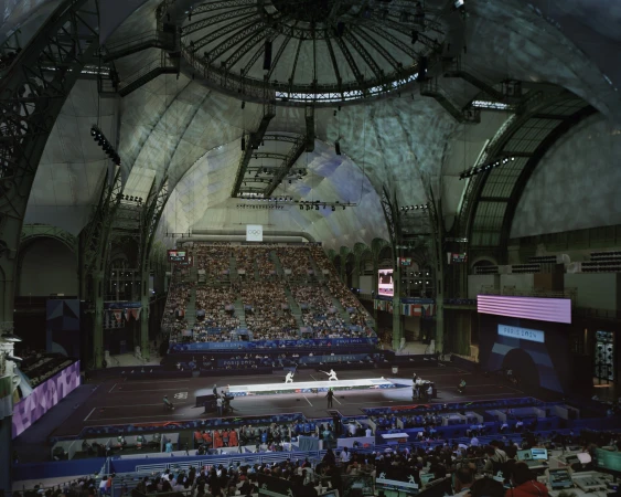 Indoor fencing competition inside an ornate event space filled with spectators, fencing pistes, flags, and large video screens.