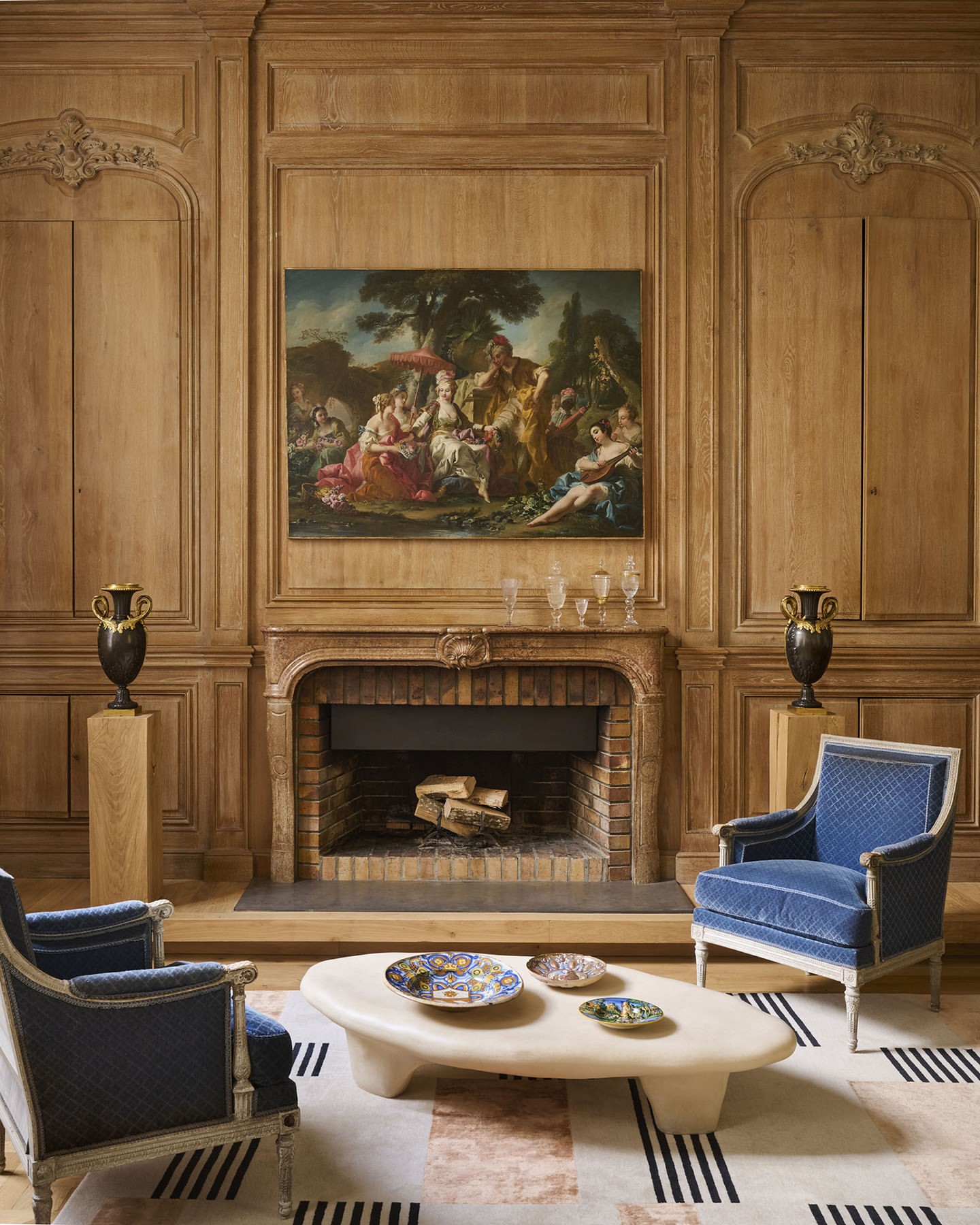 Living room interior with a fireplace, painting above the mantel, two blue upholstered chairs, and a geometric pattern rug on the floor.
