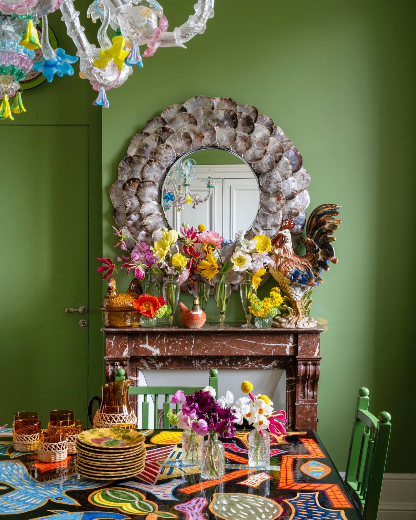 Eclectic dining room featuring an array of colorful flowers in vases, vintage plates, and a decorative rooster sculpture above the marble fireplace.
