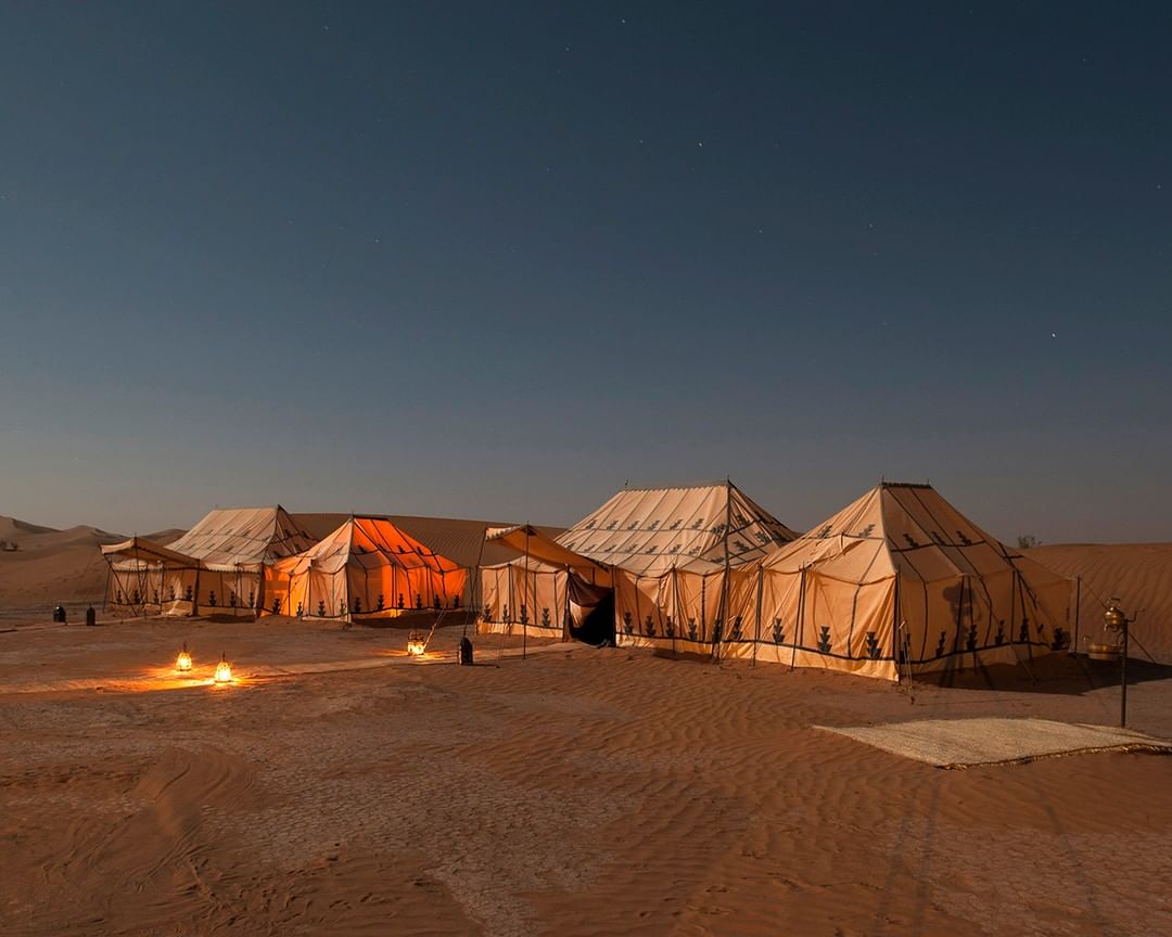 Desert camp at night with illuminated tents. Clear dark sky. Desert landscape. Low angle. Earthy tones. Rug on the ground.