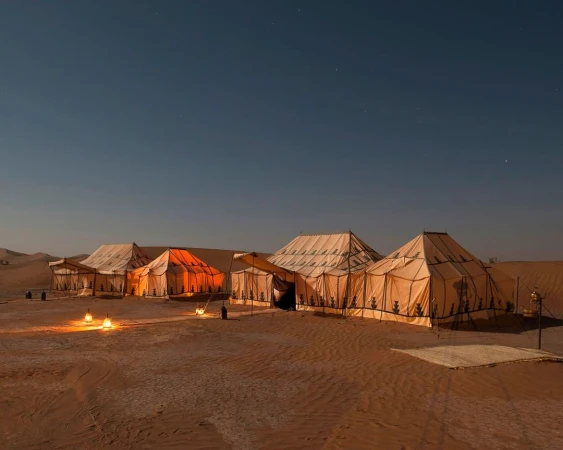 Desert camp at night with illuminated tents. Clear dark sky. Desert landscape. Low angle. Earthy tones. Rug on the ground.