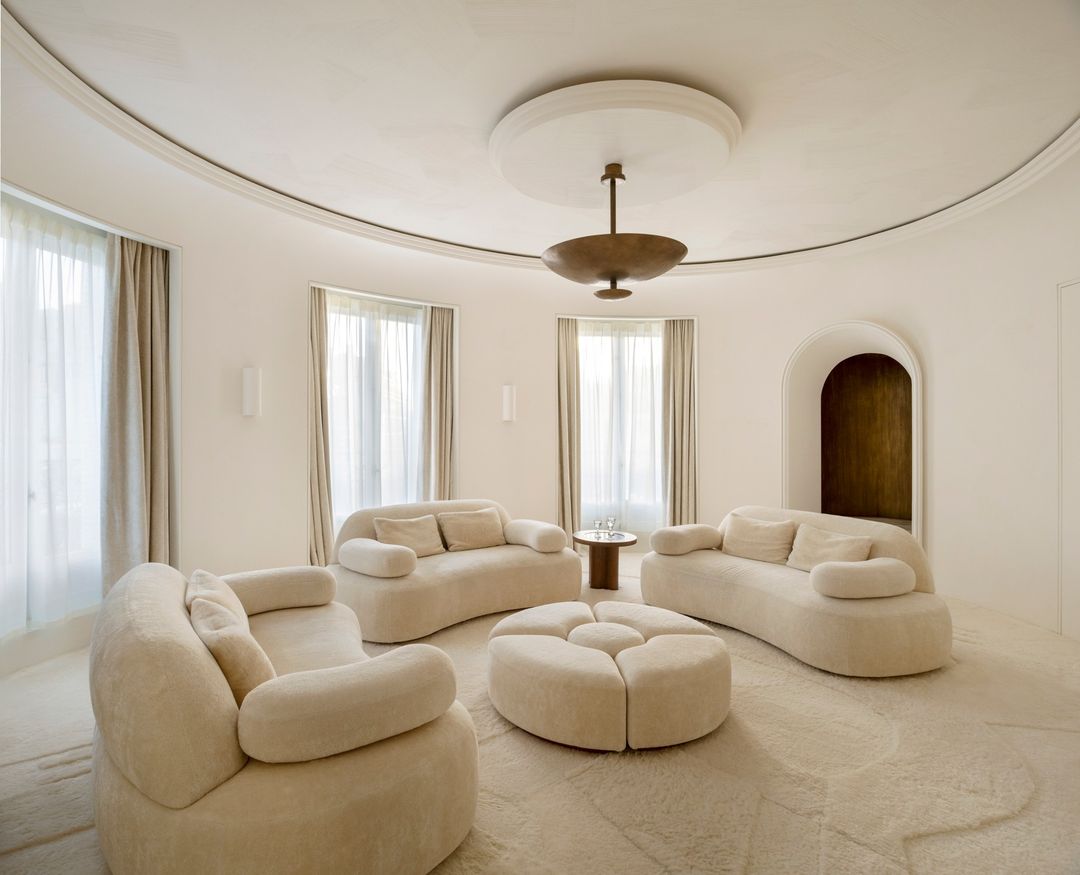 Bright, light-filled interior featuring curved sofas, round ottoman, and elegant decor with neutral colors and natural light from windows.