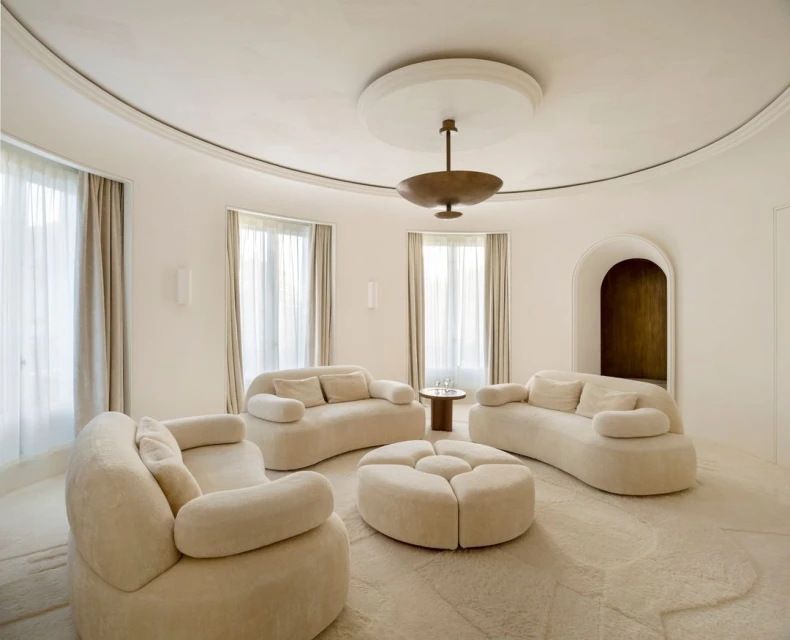 Bright, light-filled interior featuring curved sofas, round ottoman, and elegant decor with neutral colors and natural light from windows.