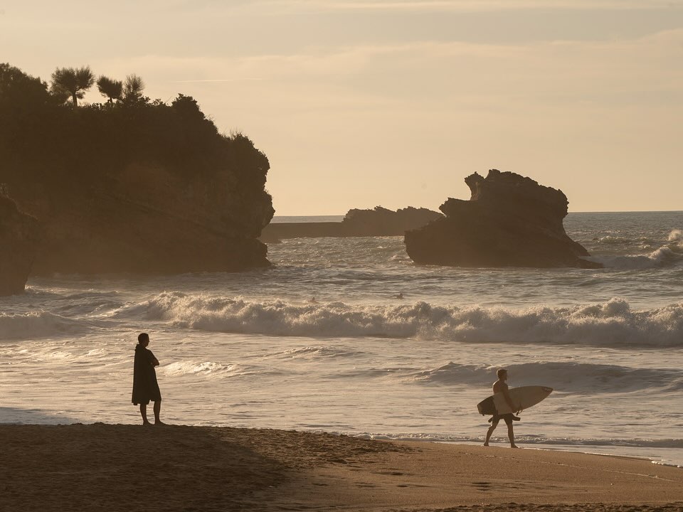 Silhouetted figures on a sandy beach with crashing ocean waves, rocky cliffs, and distant trees under an overcast sky.