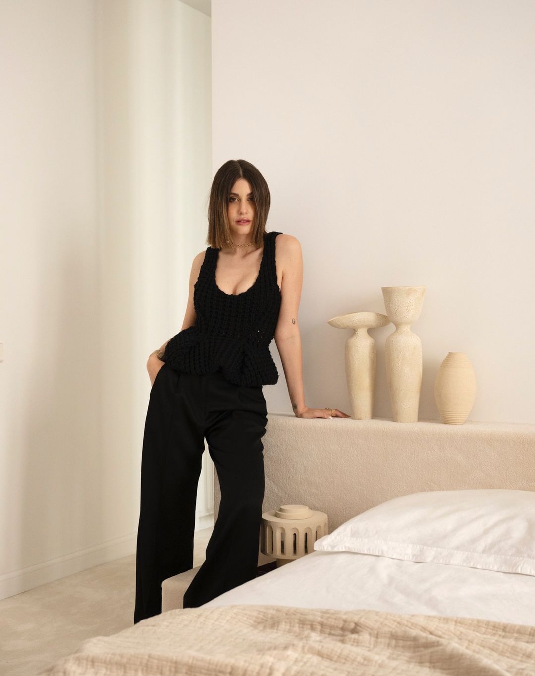 A brunette woman in a black knit top and black pants poses indoors with neutral tones, offering a stylish, minimalist aesthetic.
