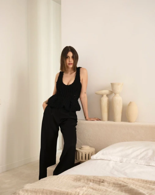 A brunette woman in a black knit top and black pants poses indoors with neutral tones, offering a stylish, minimalist aesthetic.