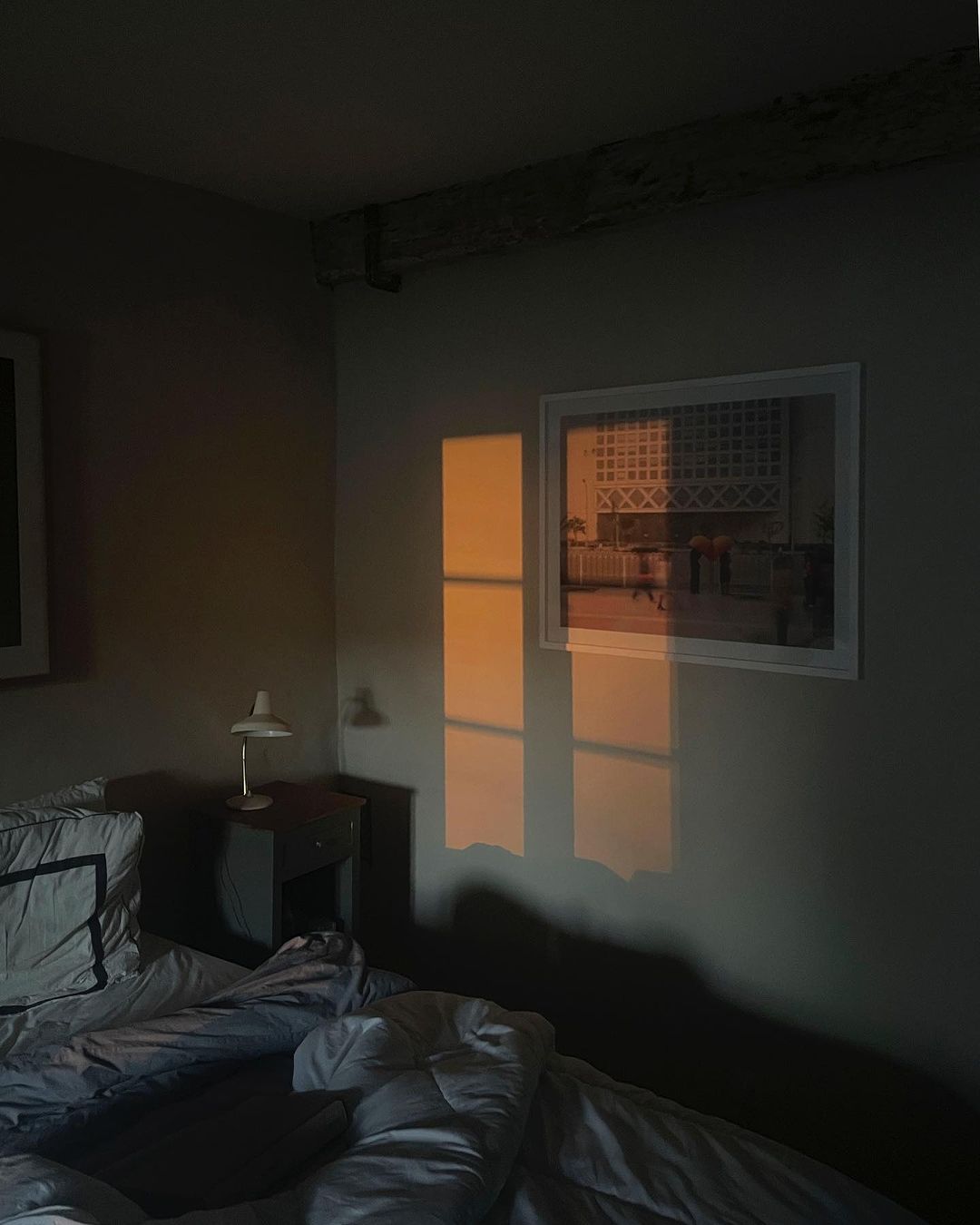 Bedroom interior with an unmade bed, nightstand and lamp, picture on the wall, and window light casting rectangular shapes on the wall.