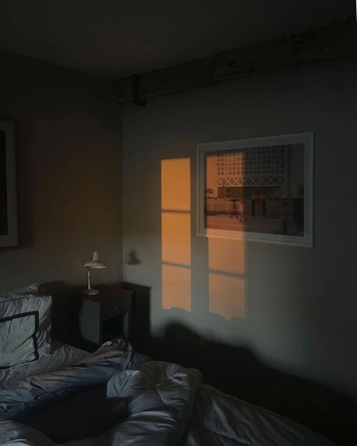 Bedroom interior with an unmade bed, nightstand and lamp, picture on the wall, and window light casting rectangular shapes on the wall.