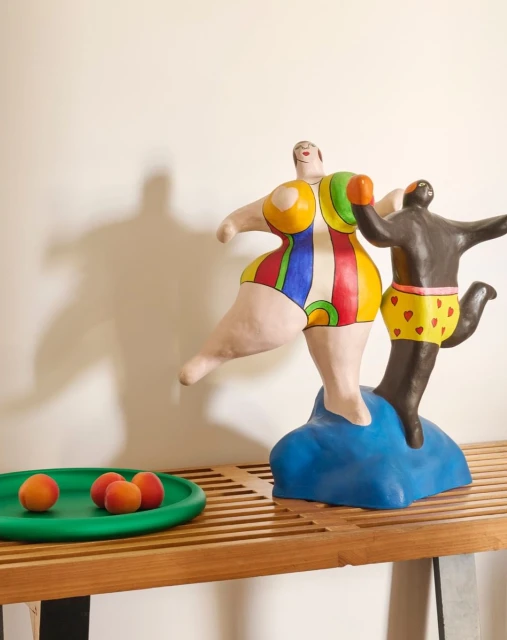 Whimsical art featuring two figures on a bench, one wearing a colorful swimsuit and the other a heart-patterned swim trunk, with peaches.