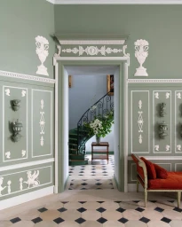 Sage green interior hallway with white neoclassical relief mouldings, tiled checkerboard floor, and a red velvet daybed.