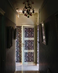 Narrow domestic hallway with dark patterned wallpaper, tiled floor, a lit chandelier, and a closed door at the end