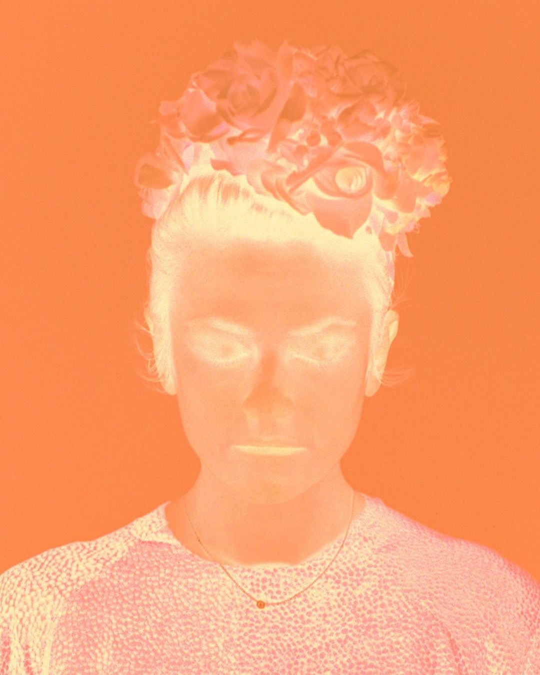 Portrait of a woman with a floral headpiece against an orange background. The image is inverted, creating a high-contrast, unique visual effect.