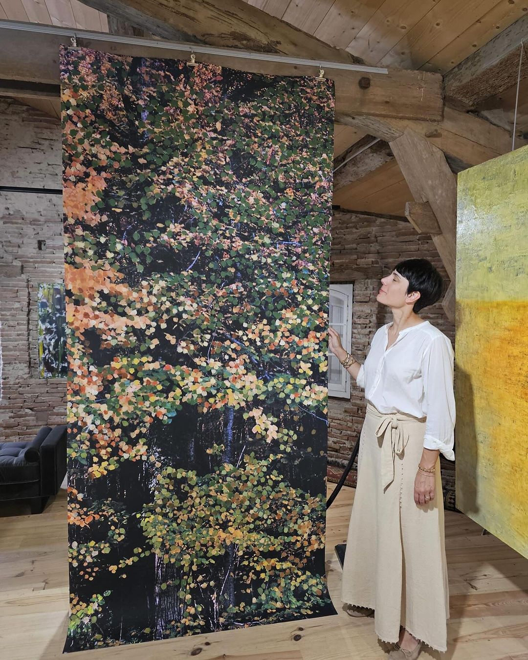 Indoor shot of an adult woman standing next to a vertical art installation with a nature scene, in a room with wood beams.
