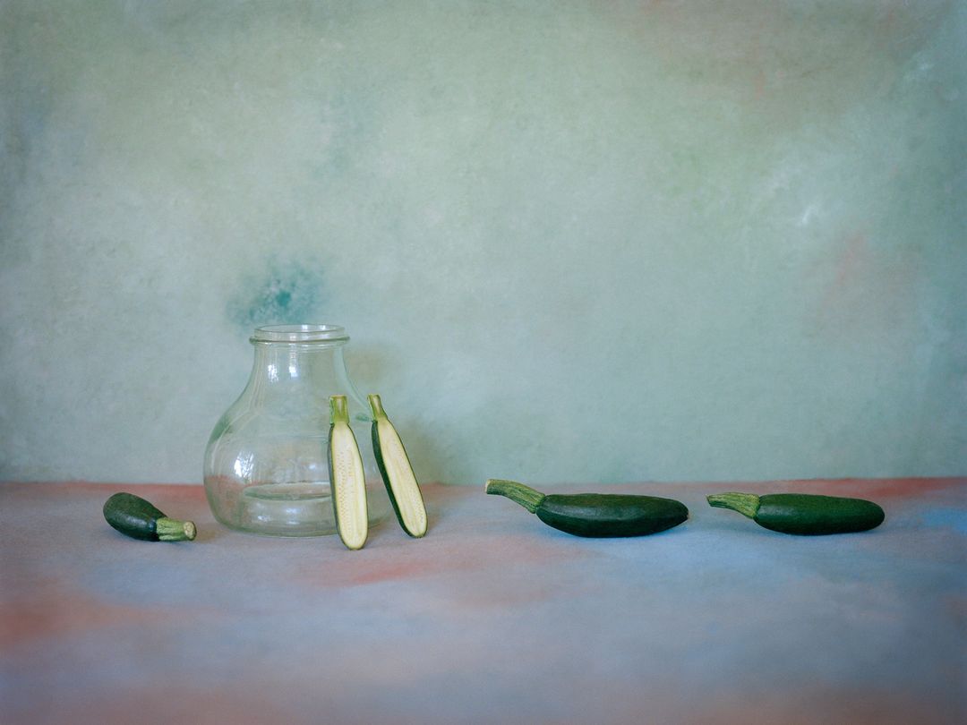 Still life featuring green zucchini squash, halved zucchini, and a clear glass vase against a mottled green background.