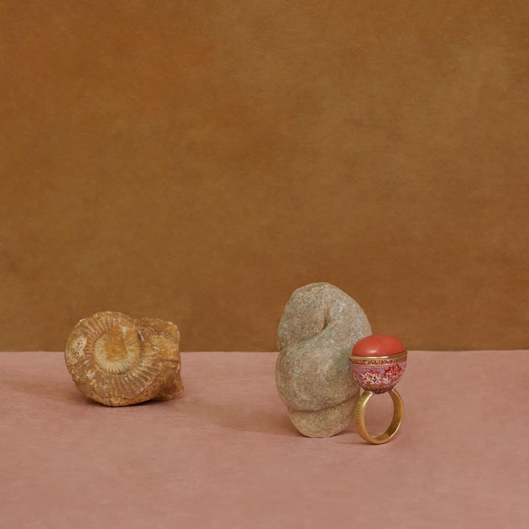 Ring with coral-colored top on a pink surface, next to a grey stone and a fossil on a pink surface against a brown background.