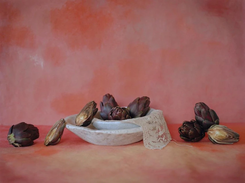 Artichokes are arranged in a white bowl, with some whole and some halved, alongside a crocheted fabric on a coral surface.