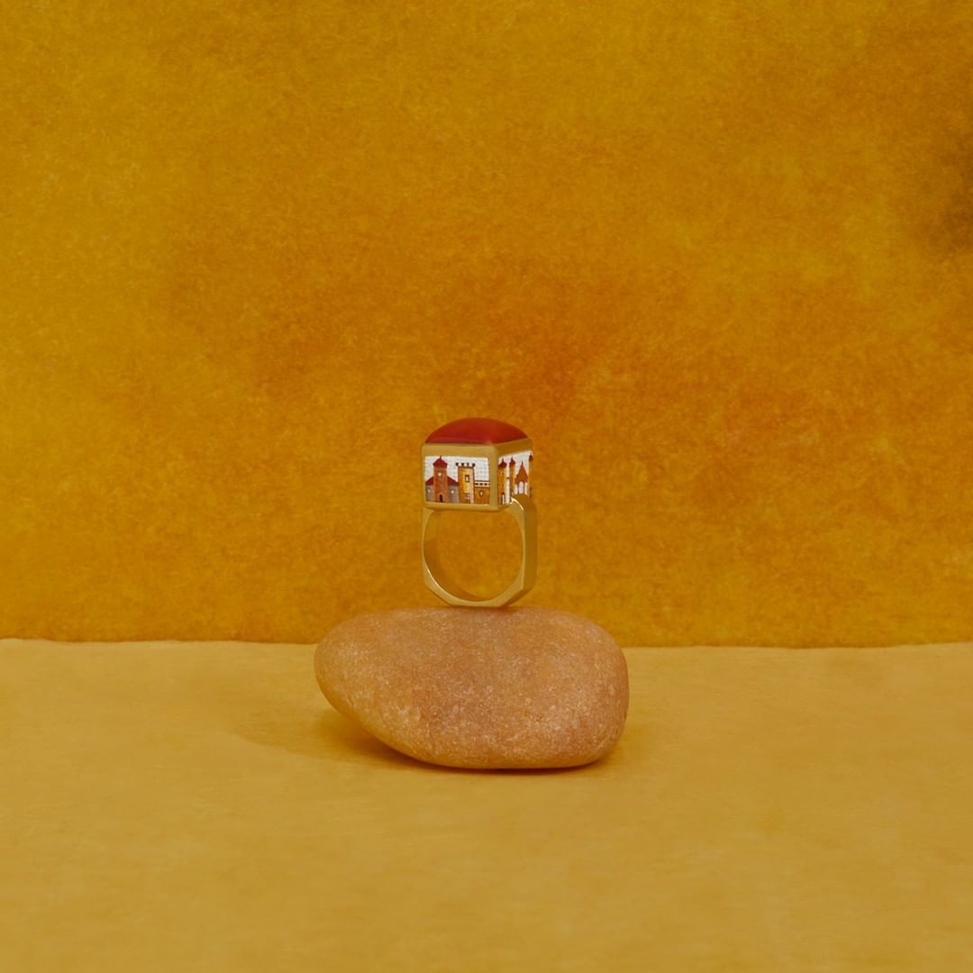 Unique ring featuring building facades and a red top, resting on a smooth stone against a textured mustard-yellow background.