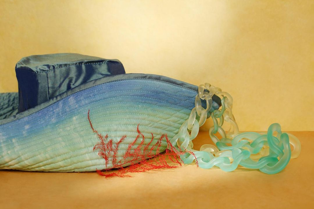 A blue bucket hat with an ombre effect and a chunky translucent chain, resting on a beige surface against a soft yellow backdrop.