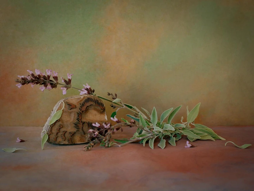 Still life photograph featuring purple sage flowers, green sage leaves, and a small decorative stone sculpture against an artistic backdrop.